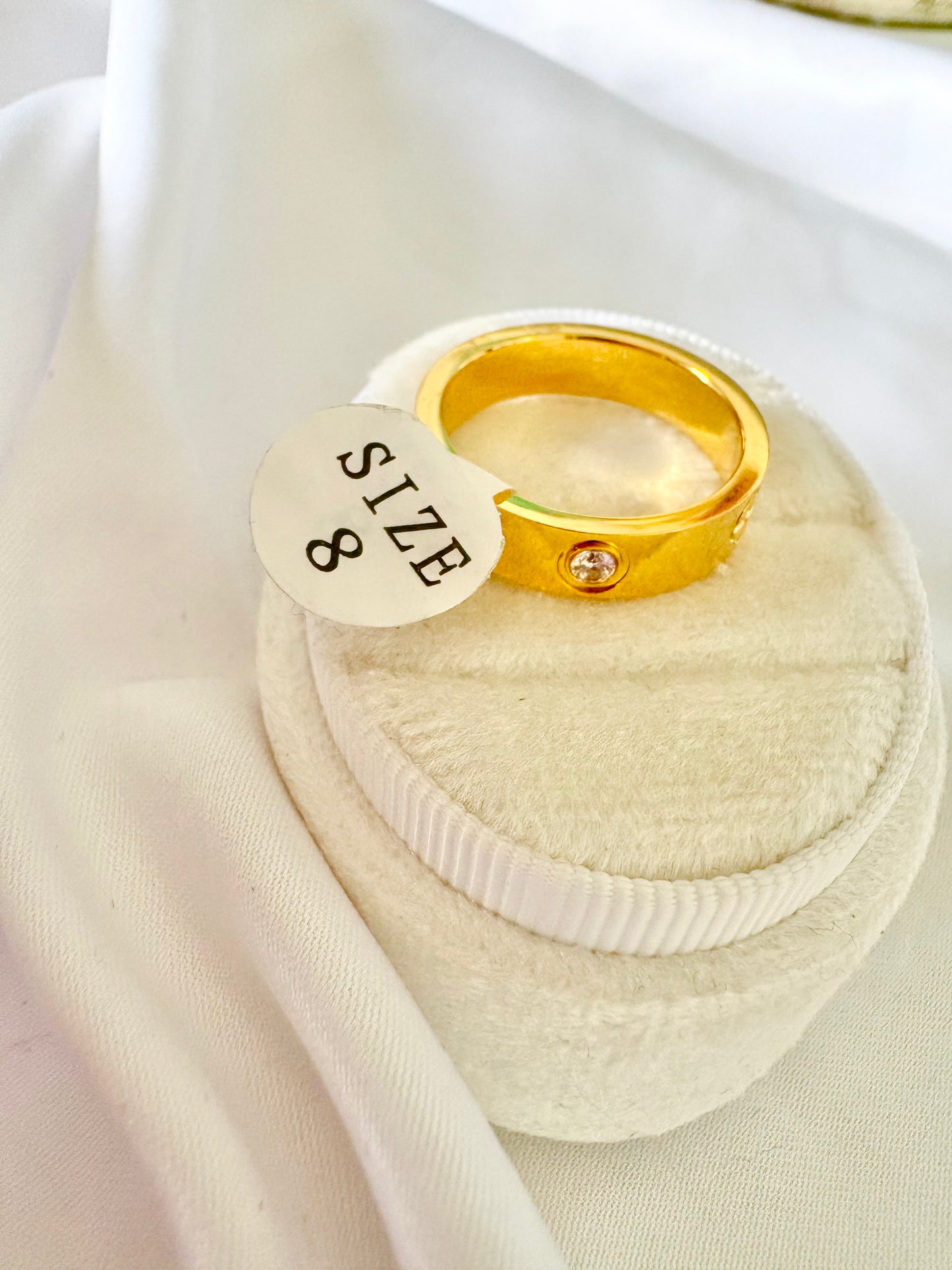 A gold band ring with a small clear gemstone, displayed on a velvet ring box 'SIZE 8'.