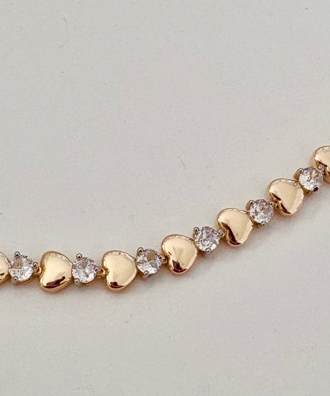 Gold bracelet with heart-shaped links and embedded zircon on a light gray background