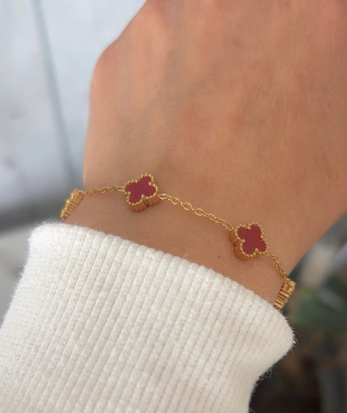 Gold bracelet with red wine clover-shaped charms on a wrist against a blurred background