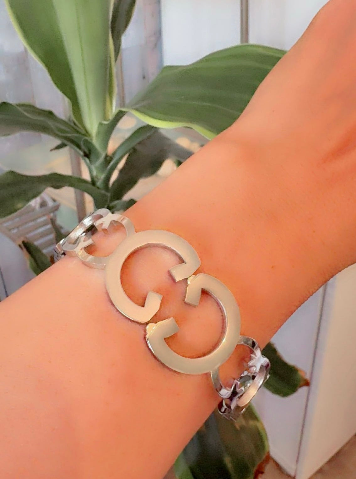 Silver bracelet with a large 'G' on a wrist, with a plant in the background