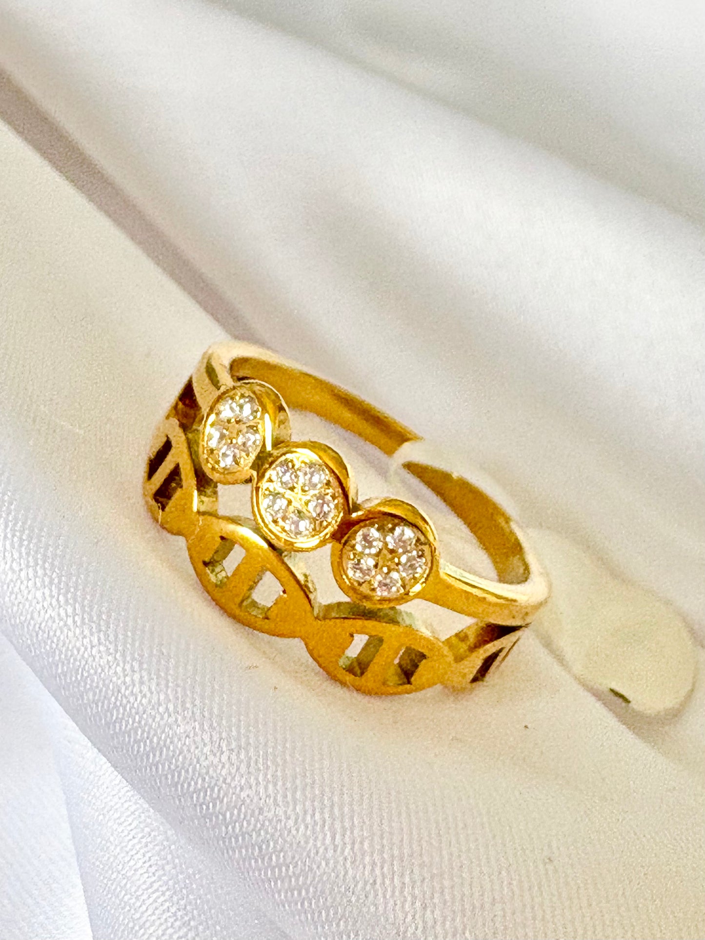 A gold ring with three oval clusters of small clear stones, displayed on a soft white fabric background.