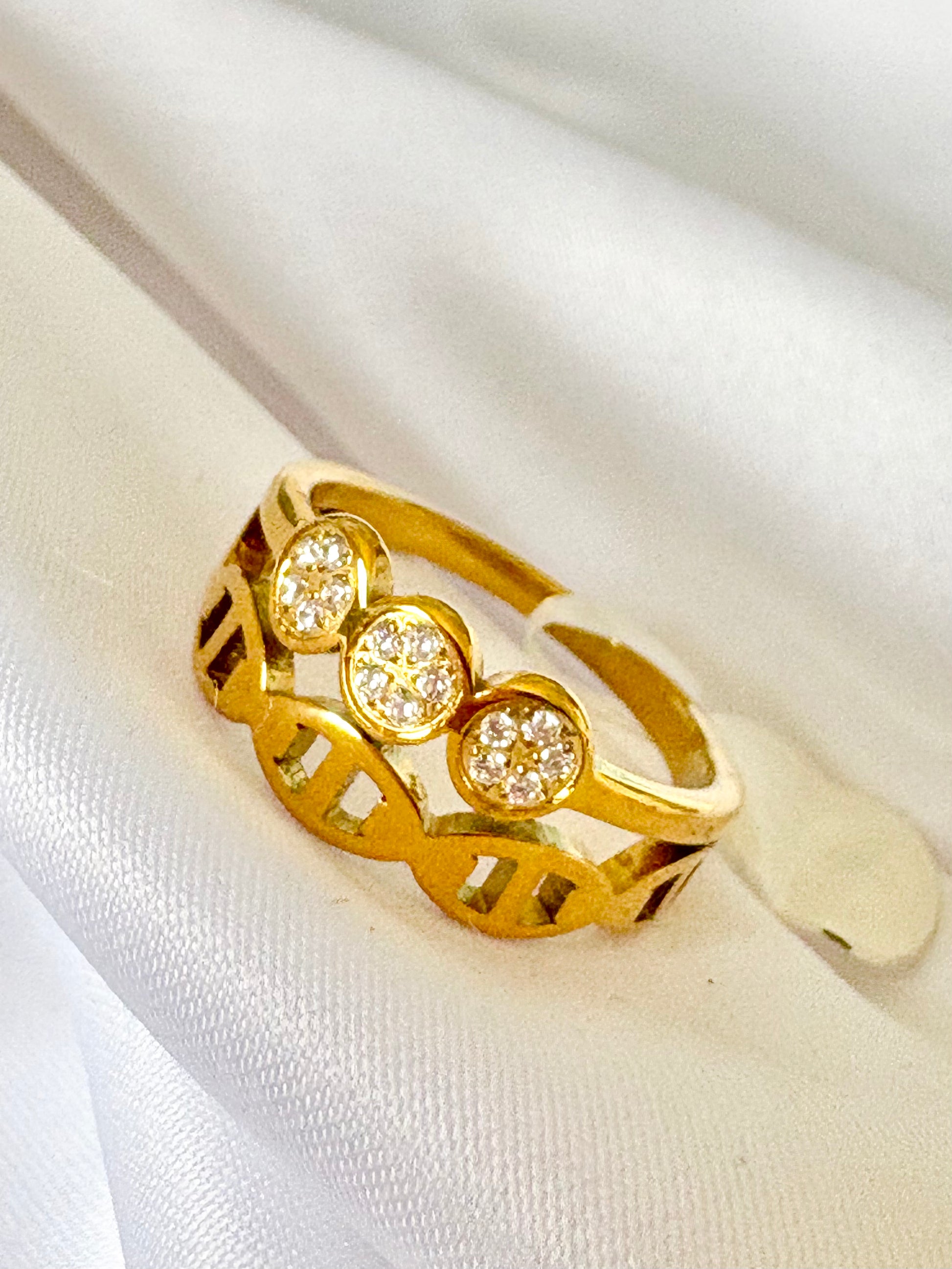 A gold ring with three oval clusters of small clear stones, displayed on a soft white fabric background.