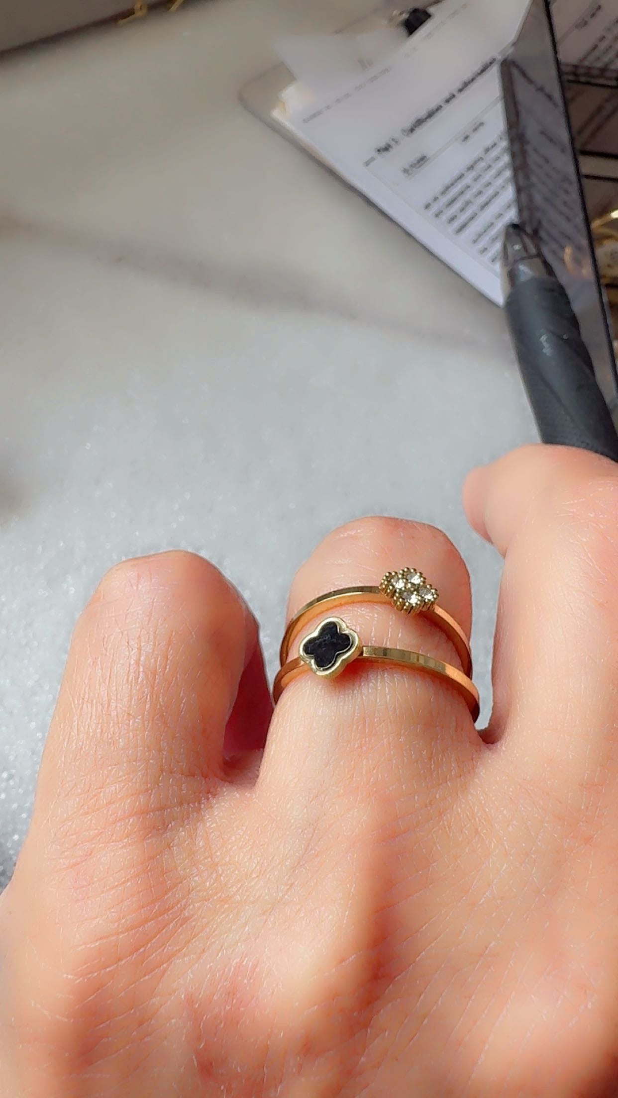 A close-up of a hand wearing two gold rings: one with a black clover motif and the other with a small floral cluster