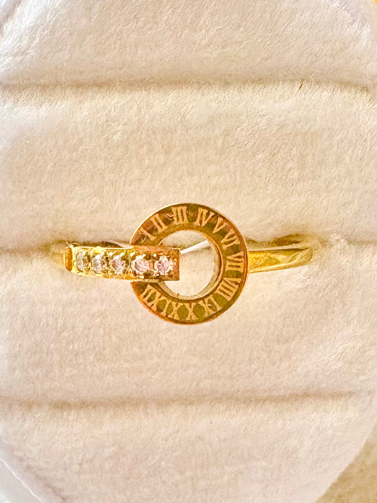 A gold ring features a circular element with Roman numerals and a band with a row of small clear stones.