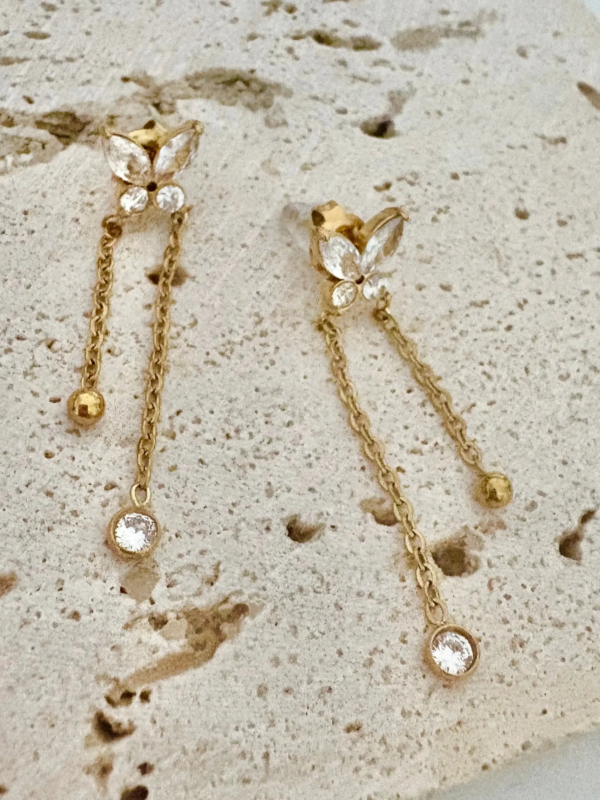 A pair of gold butterfly drop earrings with clear rhinestone accents, displayed on a textured beige stone surface.