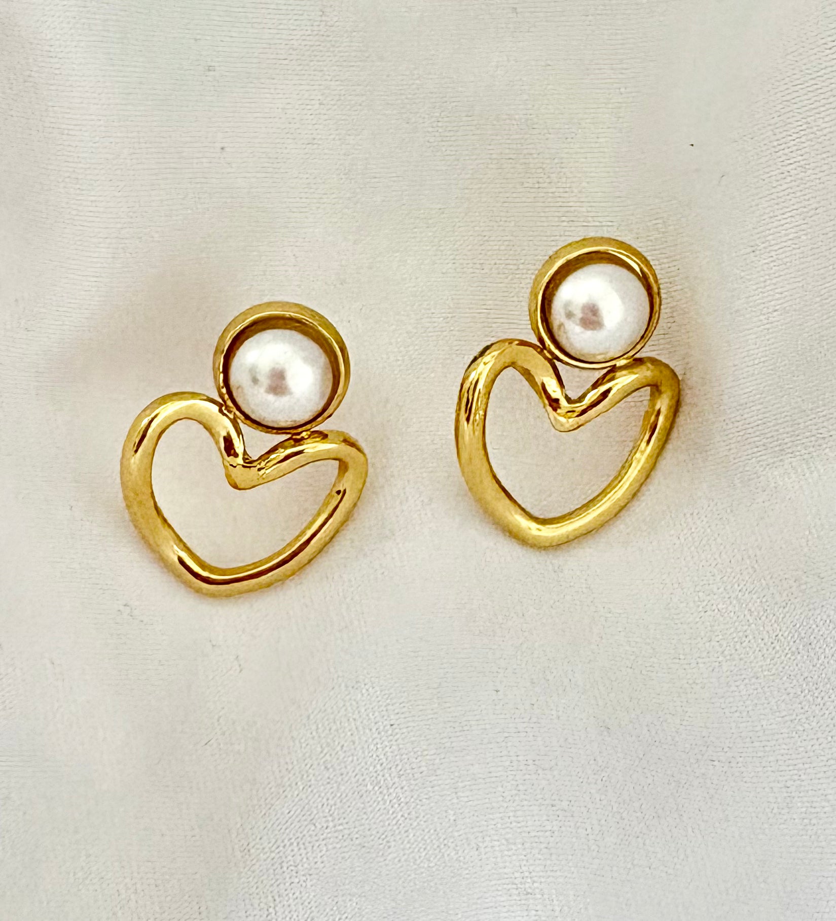 Gold heart-shaped earrings with pearl centers on a light fabric background